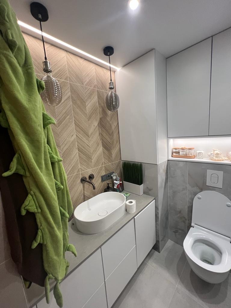 Nowoczesna łazienka z umywalką nablatową, geometryczną ścianą z płytek, wiszącą toaletą i dekoracyjnym oświetleniem, uzupełniona zielonym ręcznikiem z motywem krokodyla.