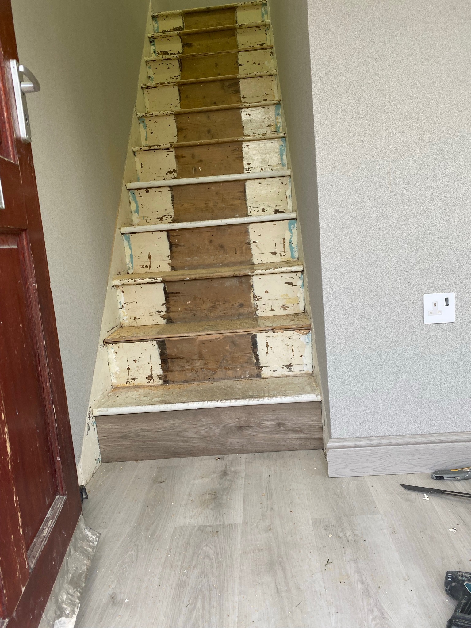 Schody drewniane w trakcie remontu: stare, częściowo zdarte stopnie z resztkami farby, nowe panele podłogowe na dole, ściana z tapetą i gniazdko elektryczne. Widoczne narzędzia na podłodze.