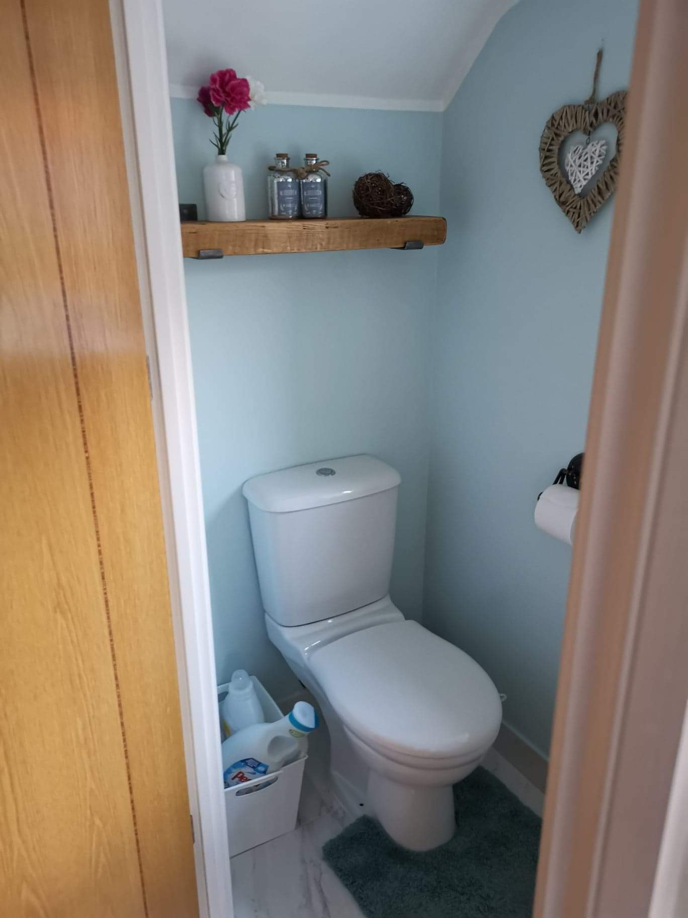 Toaleta w małej łazience z błękitnymi ścianami, drewnianą półką z dekoracjami i ozdobnym sercem na ścianie, widoczna zza częściowo otwartych drzwi.