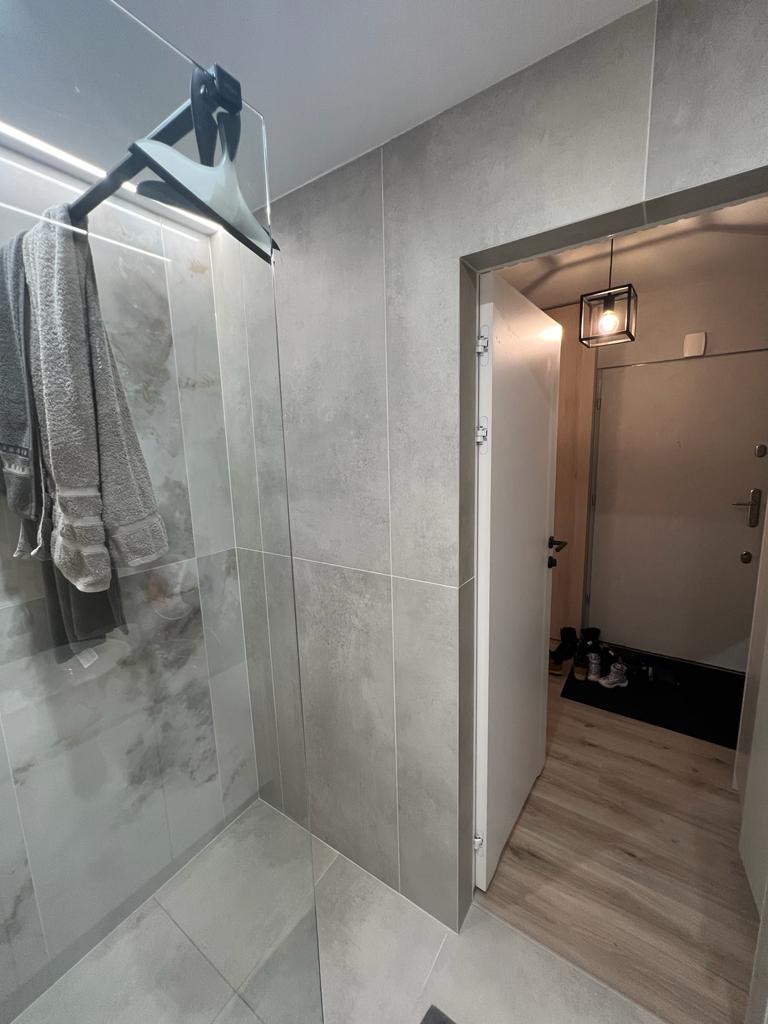 Nowoczesna łazienka z szarymi płytkami imitującymi marmur, kabina prysznicowa ze szklaną ścianką i wieszakiem na ręczniki, widok na przedpokój z drewnianą podłogą i wiszącą lampą.