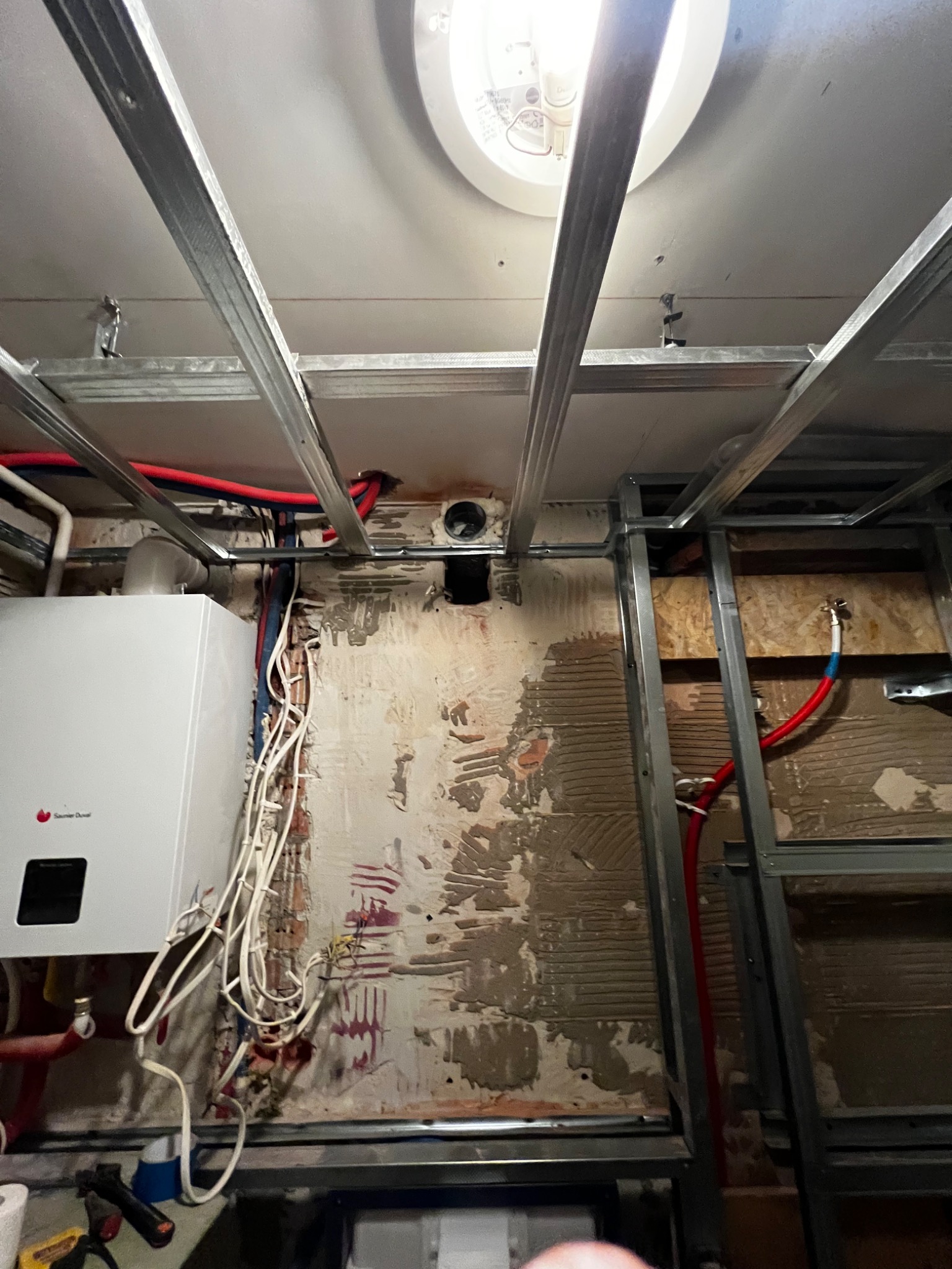 Montaż instalacji centralnego ogrzewania z kotłem gazowym na tle surowej ściany z widocznymi śladami po poprzednich pracach, konstrukcja sufitu podwieszanego w trakcie montażu.