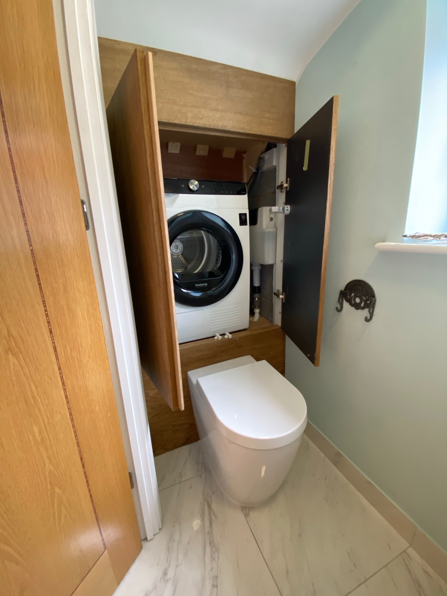 Toaleta z zabudowaną pralko-suszarką; drewniane szafki z czarnymi frontami, ceramiczna toaleta wisząca, marmurowe płytki podłogowe, ściany w odcieniach pastelowej zieleni.