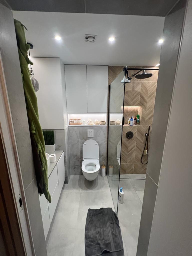 Nowoczesna łazienka z kabiną prysznicową typu walk-in, toaletą podwieszaną i szarymi płytkami imitującymi marmur, oświetlona punktowymi lampami sufitowymi, z ręcznikiem na podłodze.