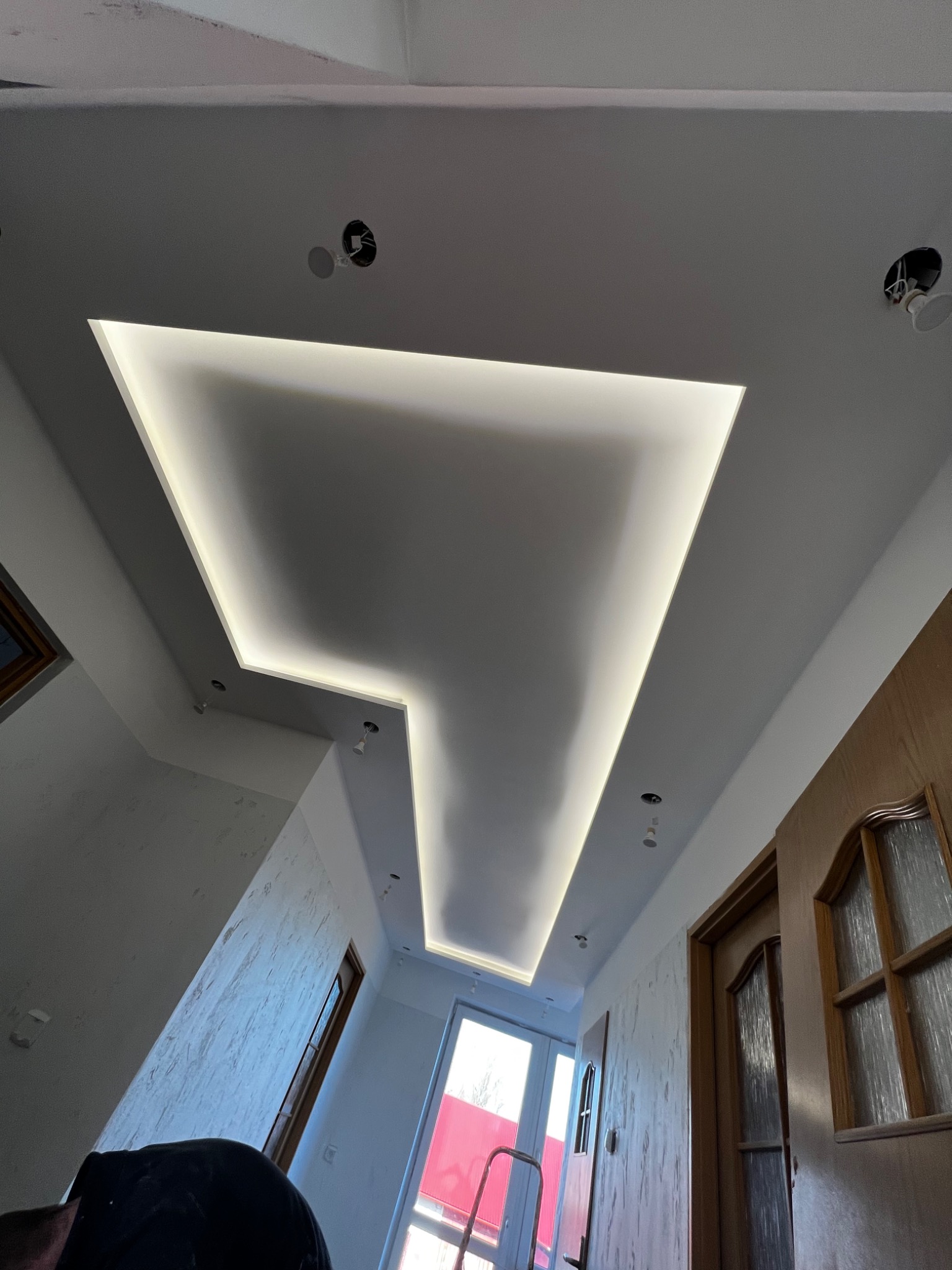 Nowoczesny sufit podwieszany z oświetleniem LED w kształcie litery L, z widocznymi otworami na oświetlenie punktowe, białe ściany z fakturą, drzwi drewniane z przeszkleniami