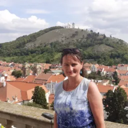 Kobieta w niebieskiej sukience pozuje na kamiennym balkonie z widokiem na miasto z czerwonymi dachami i wzgórze z białym kościołem na szczycie.