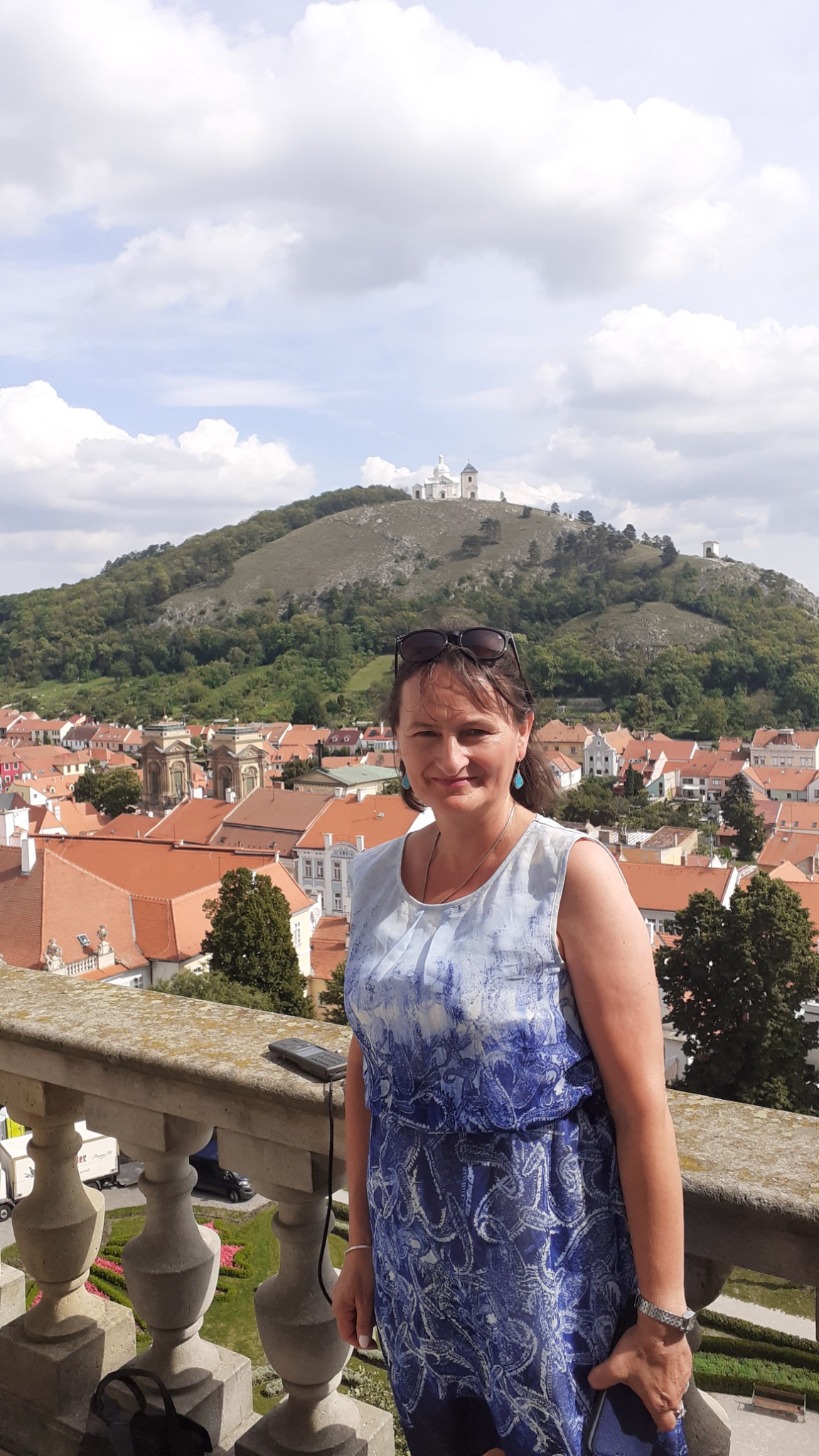 Kobieta w niebieskiej sukience pozuje na kamiennym balkonie z widokiem na miasto z czerwonymi dachami i wzgórze z białym kościołem na szczycie.