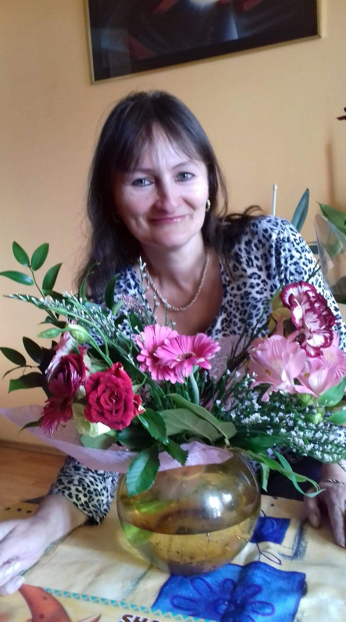 Uśmiechnięta kobieta w bluzce w panterkę pozuje z bukietem różowych i czerwonych kwiatów w okrągłym, szklanym wazonie na wzorzystym obrusie.