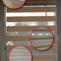 Zasłonięte okno z roletami dzień-noc w beżowo-białej kolorystyce, widoczne detale mechanizmu i konstrukcji, z zewnątrz widok na zieleń.