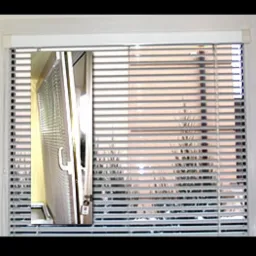 Zamknięte, poziome żaluzje aluminiowe w białej ramie okiennej, z widocznym odbiciem w szybie. W tle rozmyty widok na budynek i rośliny.