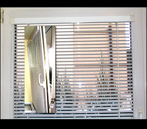 Zamknięte, poziome żaluzje aluminiowe w białej ramie okiennej, z widocznym odbiciem w szybie. W tle rozmyty widok na budynek i rośliny.
