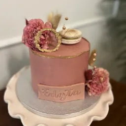 Różowy tort z dekoracją z kwiatów, złotego pierścienia i makaronika, na paterze z falbankami, z napisem 'Edyta' na froncie.