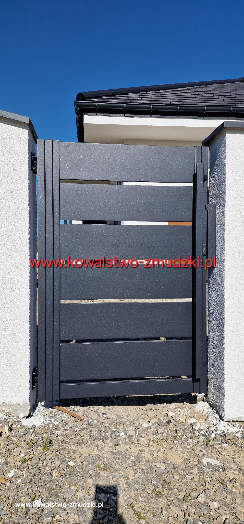 Nowoczesna, grafitowa brama wjazdowa z poziomymi szczebelkami, zamontowana między dwoma białymi słupkami. Widoczne detale montażowe i kamieniste podłoże.