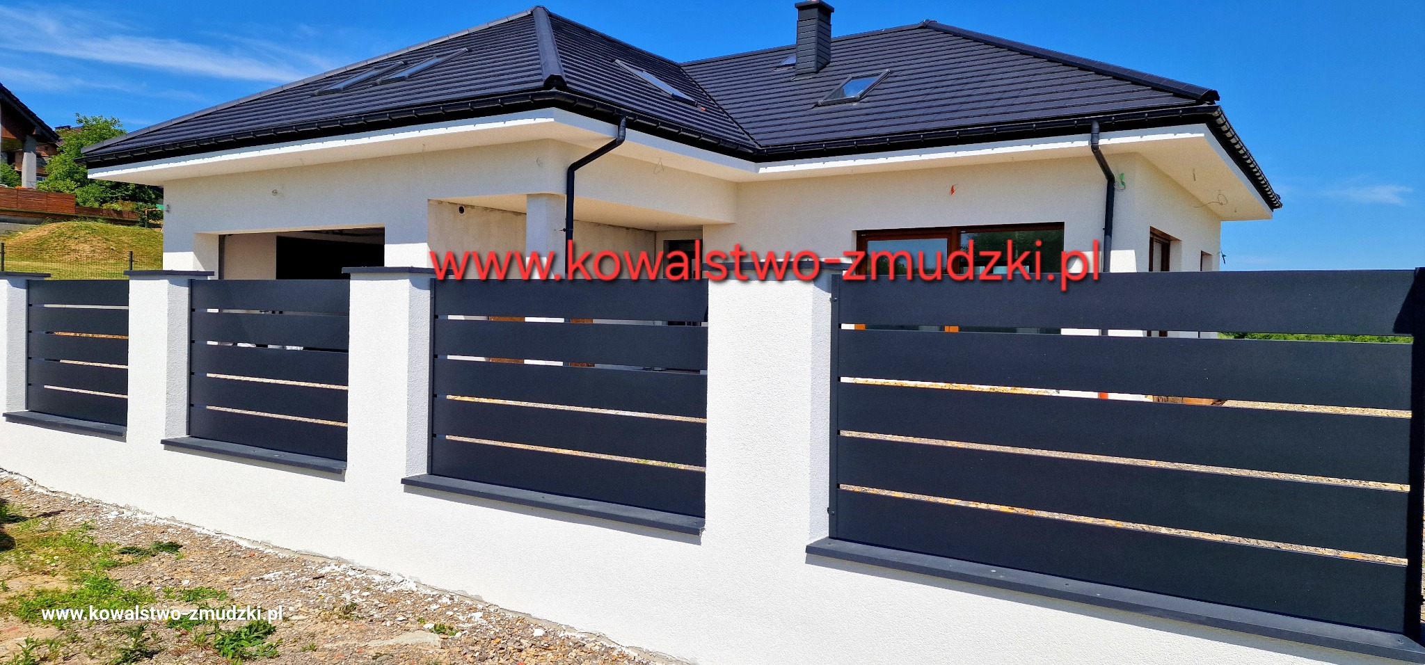 Nowoczesne, poziome przęsła ogrodzeniowe w kolorze grafitowym, osadzone na niskim, białym murku z kamienną podmurówką. W tle dom z ciemną dachówką i jasną elewacją.