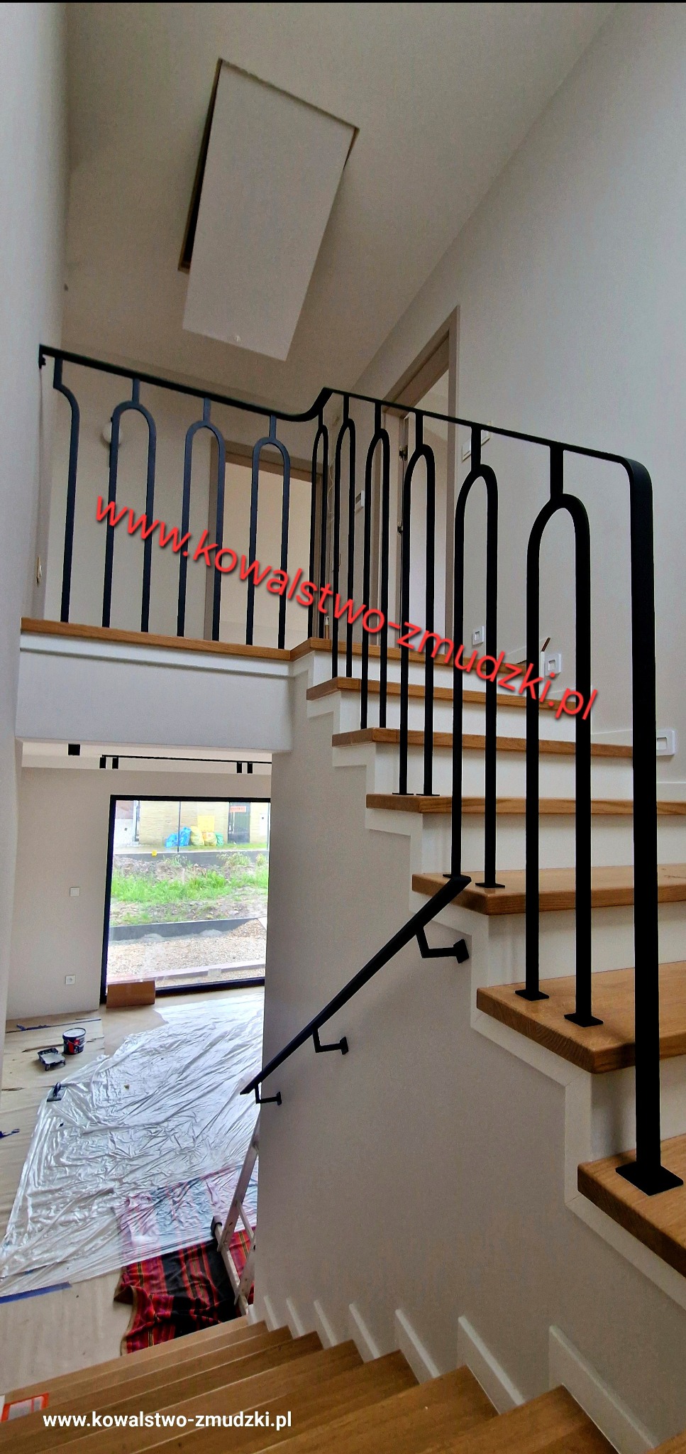 Nowoczesne, czarne poręcze schodowe z metalu, z charakterystycznymi łukami, zamontowane w jasnym wnętrzu domu. Widoczne drewniane stopnie schodów i okno w tle.