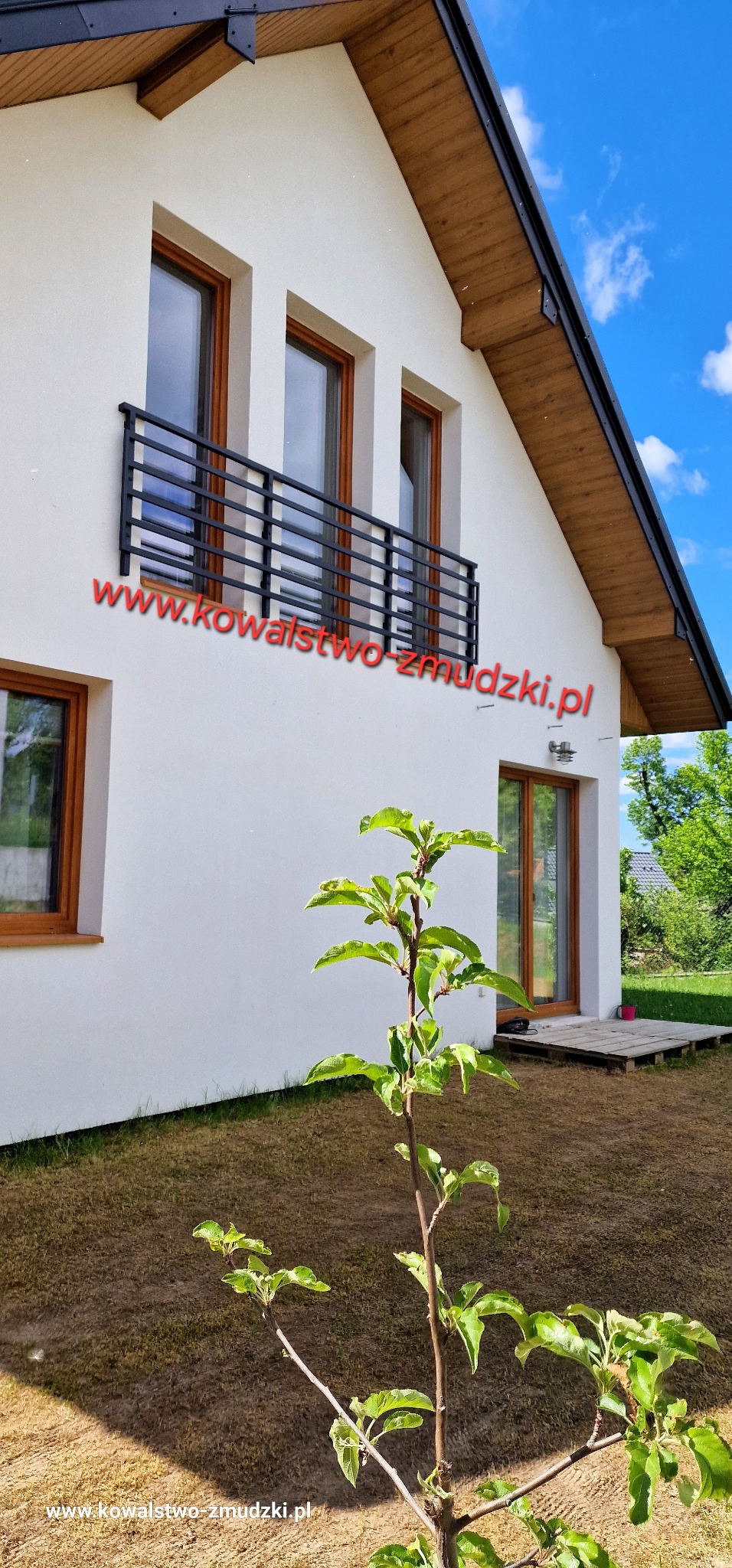 Nowoczesna, czarna balustrada balkonowa z poziomych profili na tle białej elewacji domu z drewnianą podbitką i oknami w brązowych ramach. Widok z dołu, zieleń na pierwszym planie.