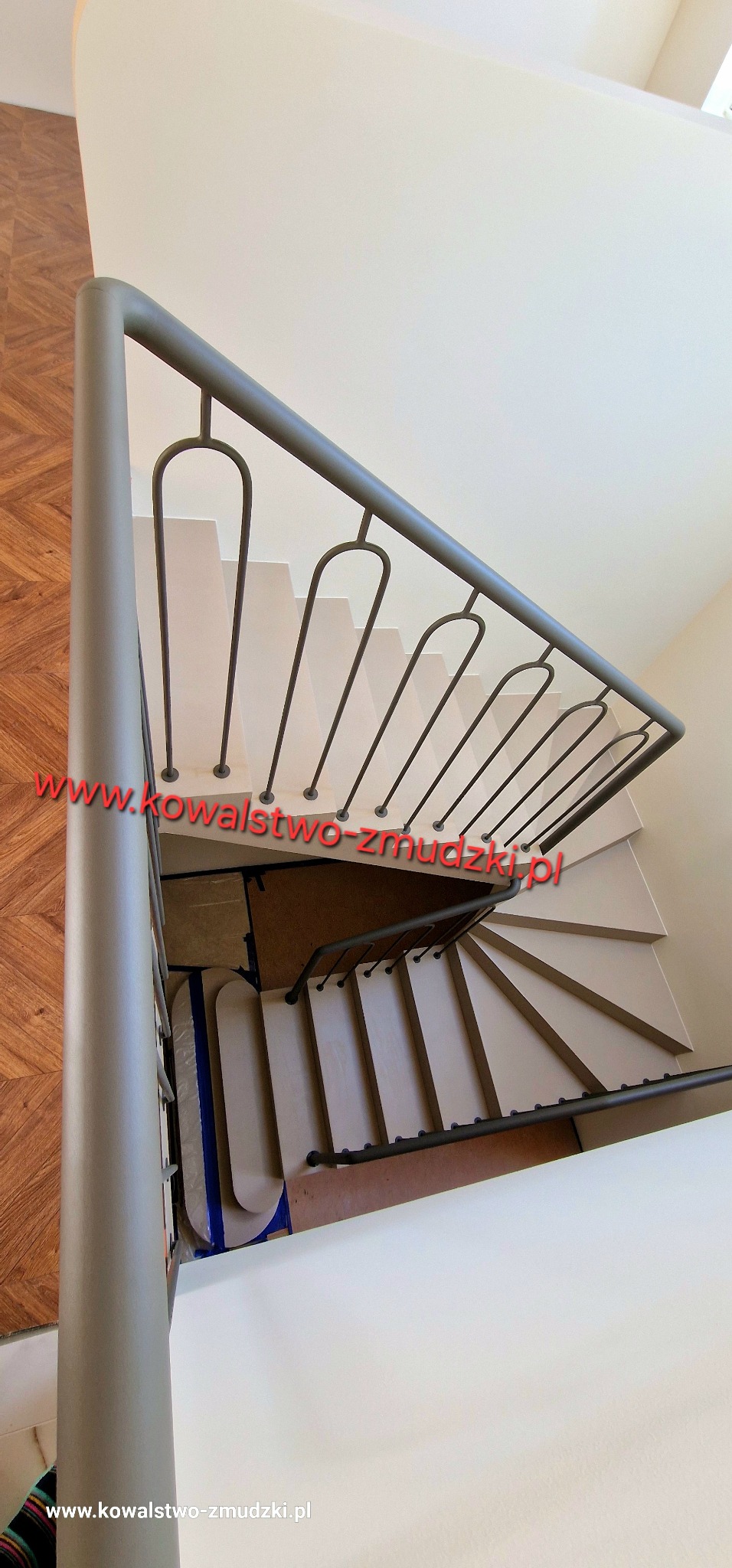 Spiralne schody z szarą, metalową poręczą, widok z góry. Nietypowy układ stopni i minimalistyczny design balustrady. Podłoga wyłożona parkietem.