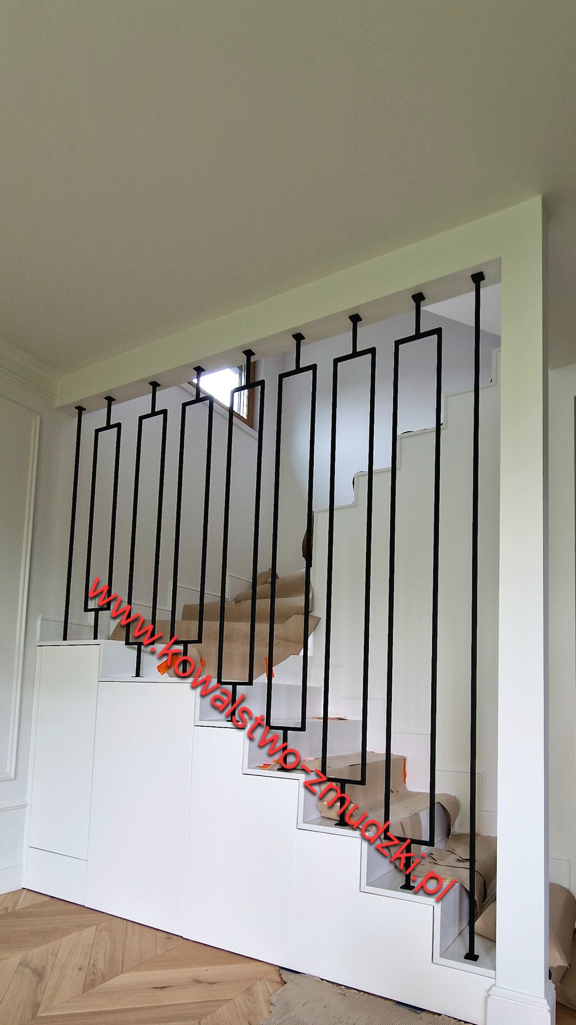 Minimalistyczna, czarna poręcz schodowa z prostokątnymi elementami, zamontowana na białych schodach z zabudową. Widoczny fragment parkietu i okno w tle.