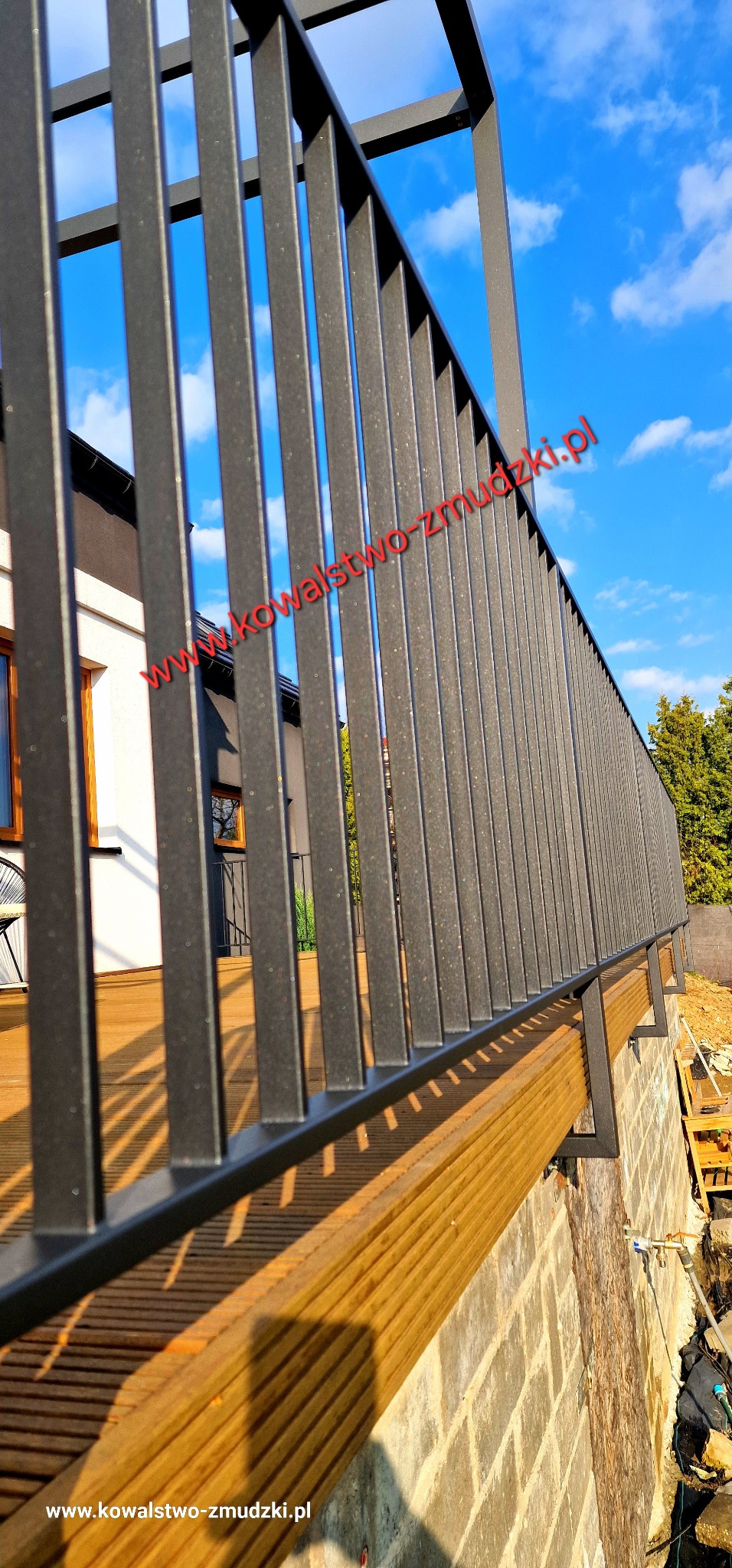 Nowoczesna balustrada metalowa w kolorze grafitowym, zamontowana na drewnianym tarasie. Widok z dołu, na tle błękitnego nieba z chmurami.