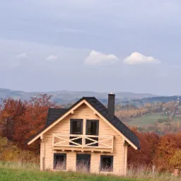 Gotowy, drewniany dom z ciemnym dachem i kominem na tle jesiennego krajobrazu z wzgórzami i lasami.