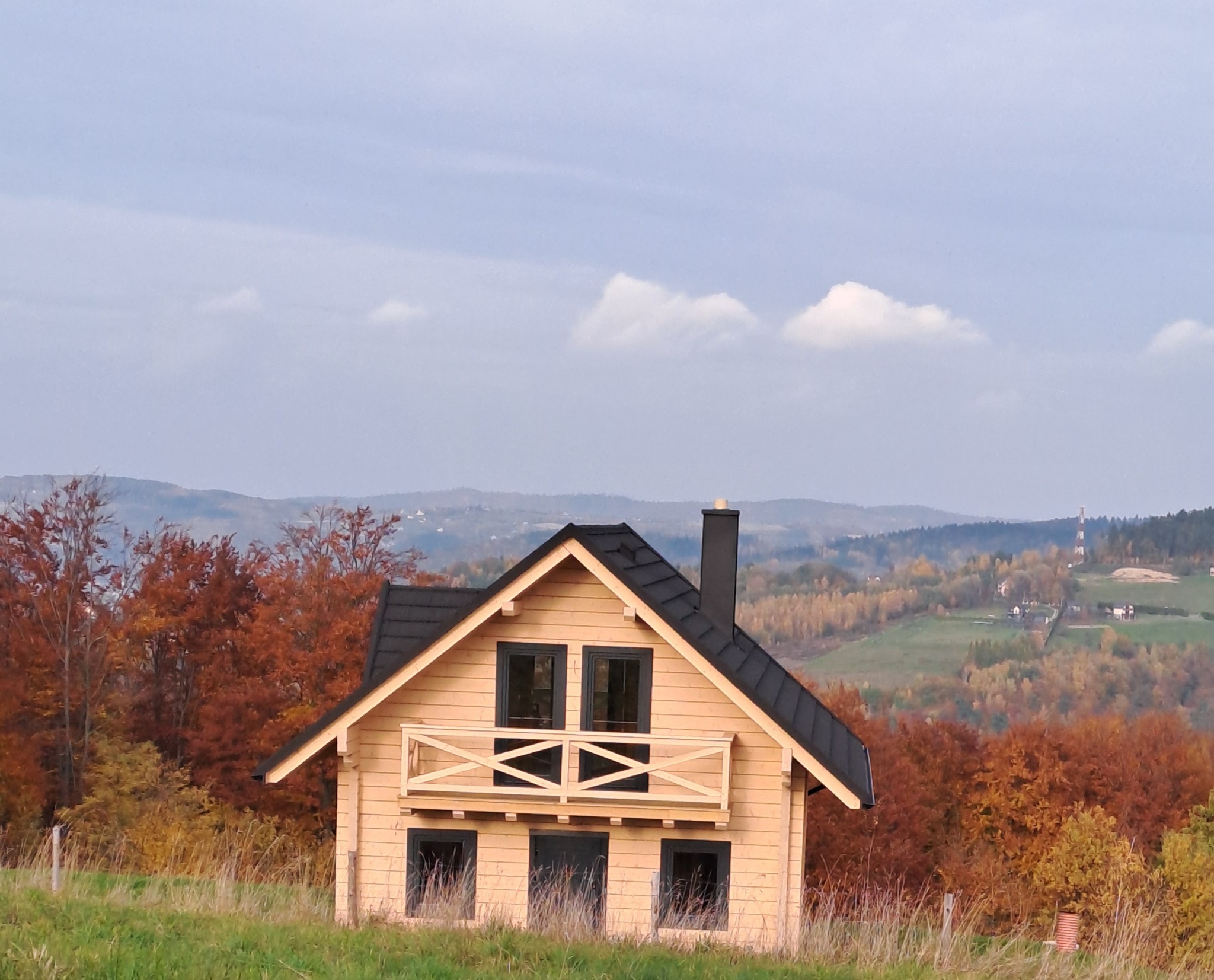 Gotowy, drewniany dom z ciemnym dachem i kominem na tle jesiennego krajobrazu z wzgórzami i lasami.