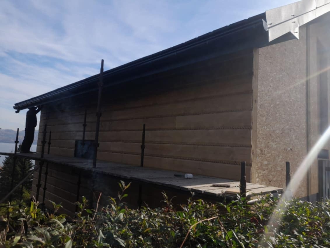 Budynek w trakcie obkładania drewnianą boazerią, widoczne rusztowanie i pracownik przy rynnie, z elementami obróbki blacharskiej.