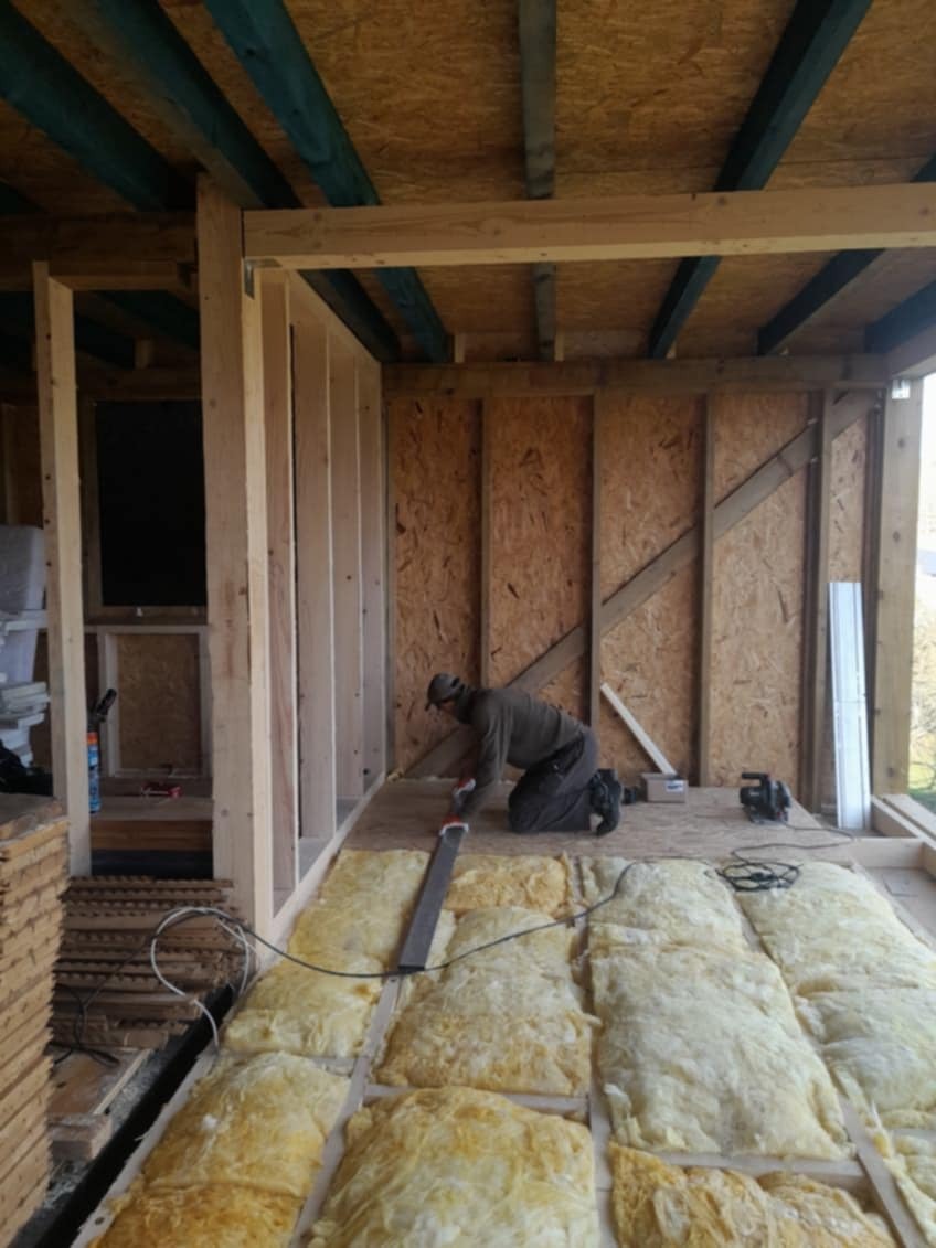 Pracownik na kolanach przykłada łatę do podłogi z izolacją z wełny mineralnej w surowym pomieszczeniu z drewnianą konstrukcją szkieletową.