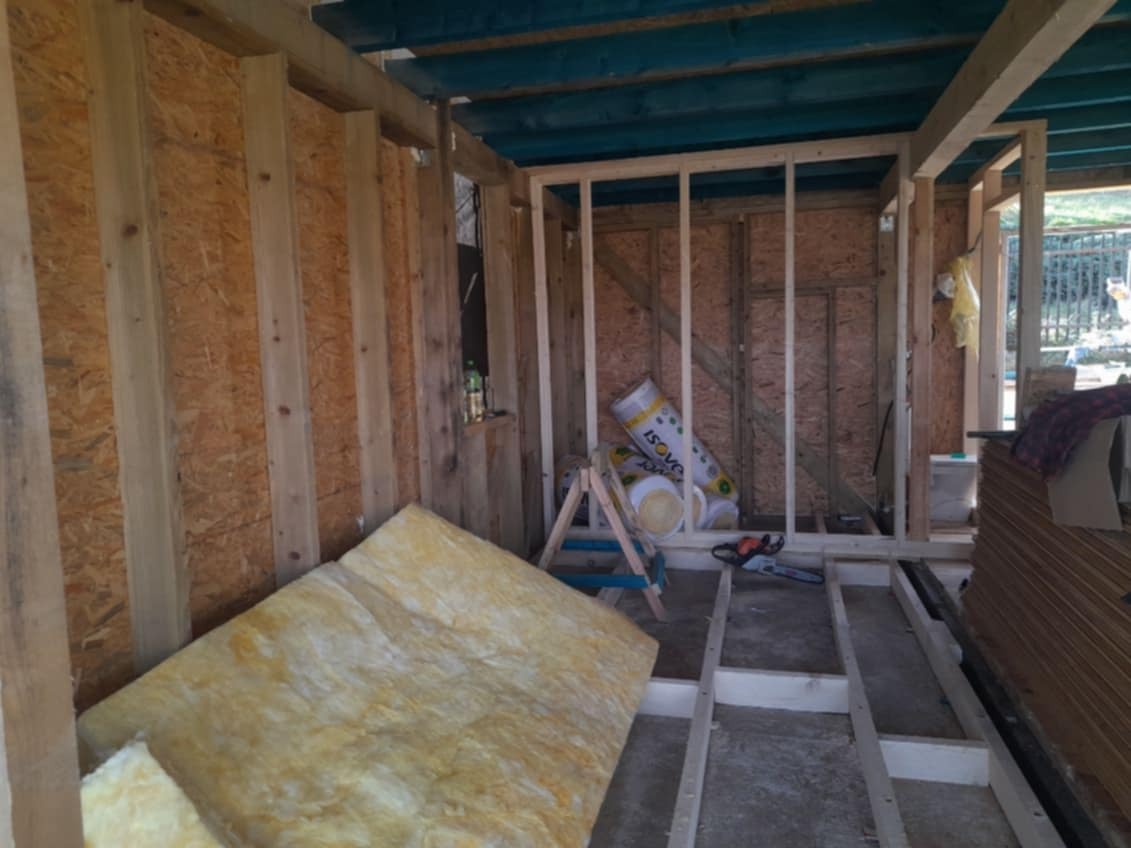 Wnętrze budynku w trakcie budowy dachu: widoczna drewniana konstrukcja więźby, płyty OSB na ścianach, izolacja termiczna ISOVER w rolkach oraz fragmenty wełny mineralnej, piła łańcuchowa, butelka...