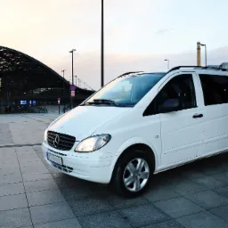 Biały Mercedes Vito z relingami dachowymi, zaparkowany przed nowoczesnym budynkiem dworca kolejowego o konstrukcji stalowej o zmierzchu.