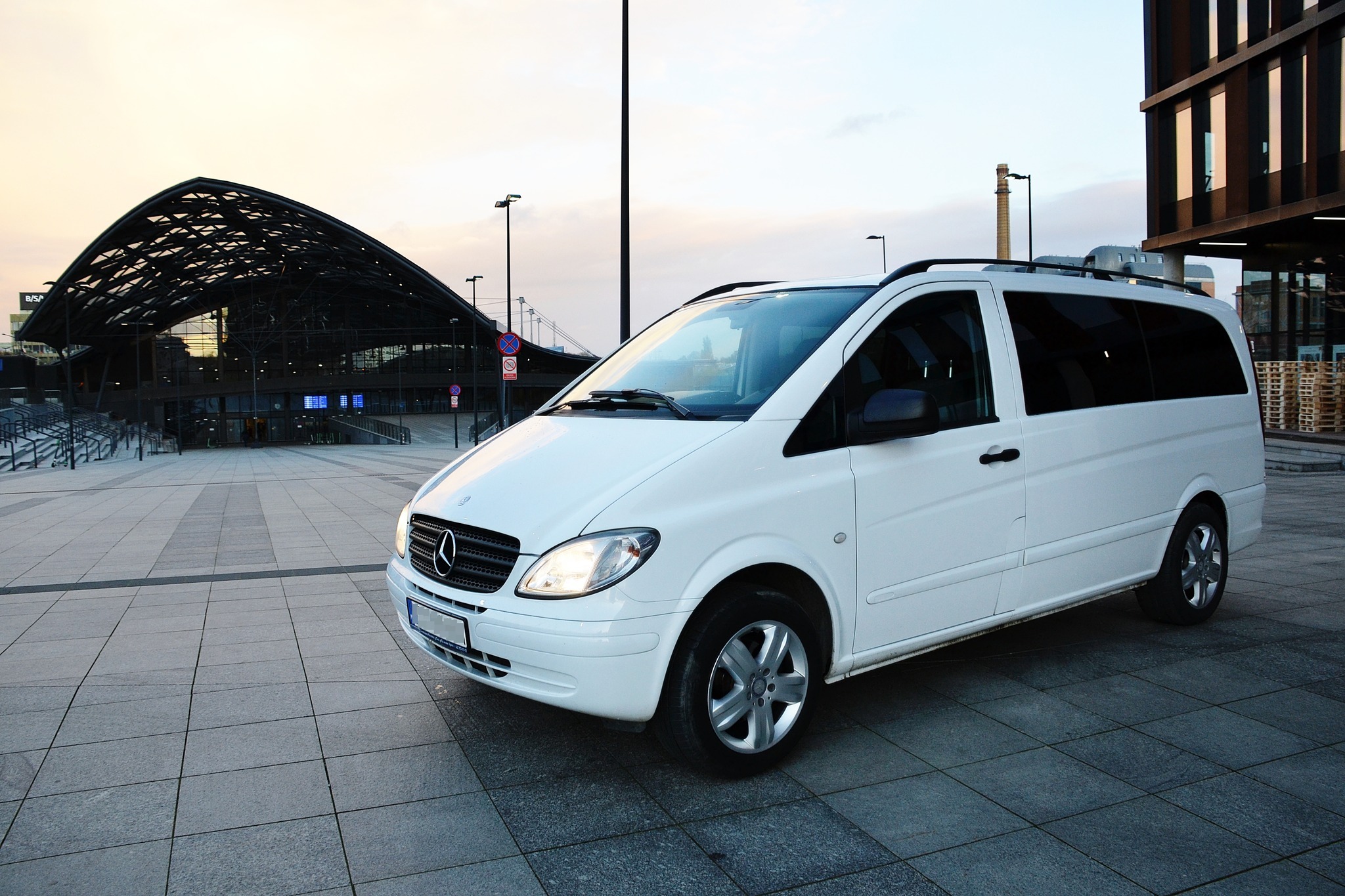 Biały Mercedes Vito z relingami dachowymi, zaparkowany przed nowoczesnym budynkiem dworca kolejowego o konstrukcji stalowej o zmierzchu.
