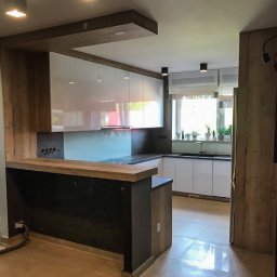 Meble Szyte na Miarę - Nowoczesna kuchnia z białymi szafkami, drewnianymi akcentami i blatem barowym z widocznymi gniazdkami elektrycznymi.