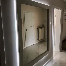 Nowoczesne lustro w ramie z podświetleniem LED, odbijające fragment przedpokoju z marmurową podłogą i białymi drzwiami.