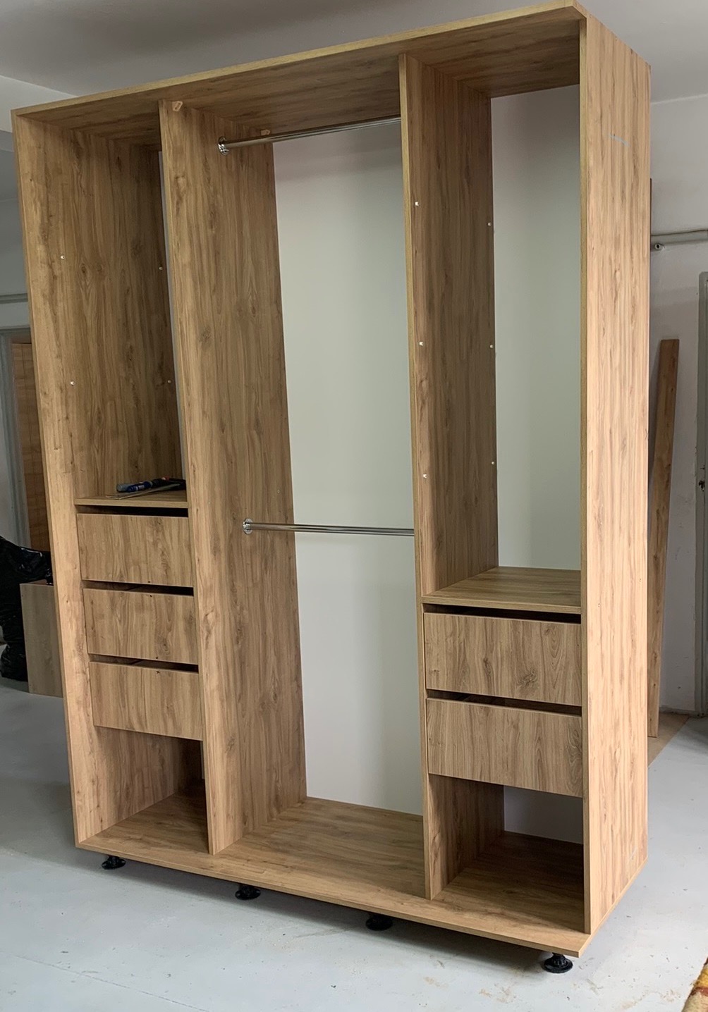 Wnętrze szafy wnękowej z jasnego drewna z szufladami i drążkami na ubrania, widoczne czarne nóżki regulacyjne, w trakcie montażu.