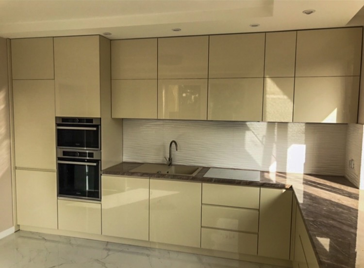 Jasna, nowoczesna kuchnia z beżowymi, lakierowanymi frontami, marmurowym blatem i wbudowanymi piekarnikami, oświetlona naturalnym światłem padającym na falistą, białą ścianę nad blatem.