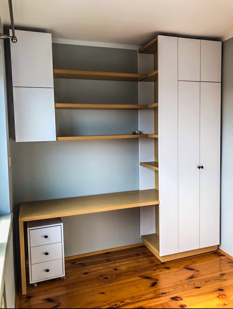 Biało-drewniane meble do pracy w rogu pokoju: szafka wisząca, biurko z kontenerkiem na kółkach i wysoka szafa z półkami.