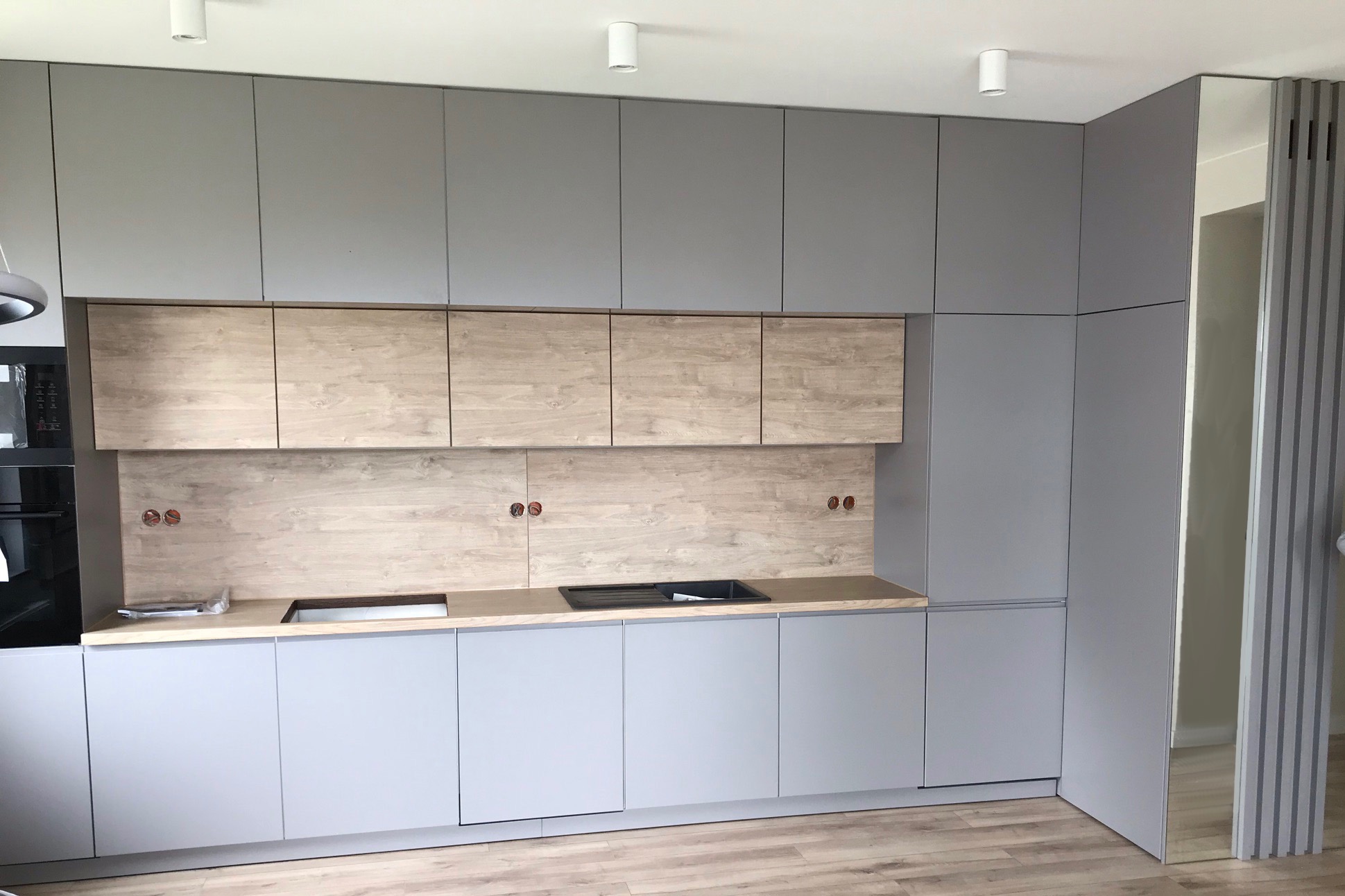 Szara zabudowa kuchenna z drewnianymi akcentami, widoczne gniazdka elektryczne w ścianie nad blatem, minimalistyczny design, podłoga imitująca drewno.