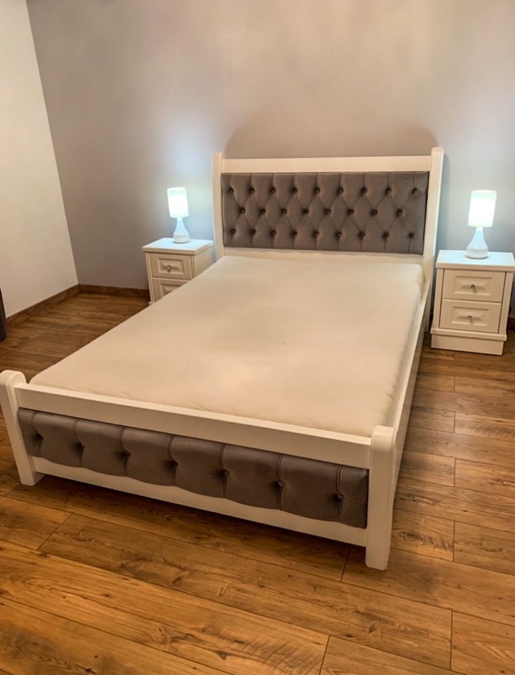 Białe łóżko z tapicerowanym, szarym zagłówkiem i bokami, uzupełnione o dwie białe szafki nocne z lampkami na drewnianej podłodze.