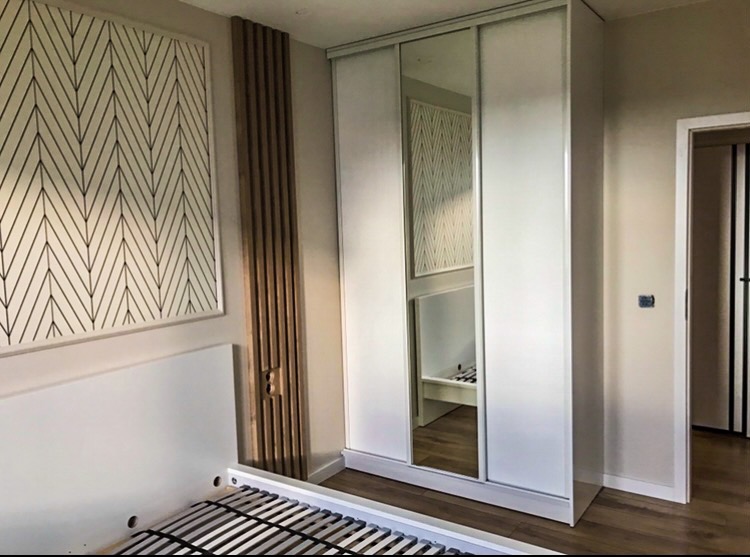 Biała szafa przesuwna z lustrem w sypialni, widoczna rama łóżka, dekoracyjny panel ścienny z pionowych listew i grafika na ścianie.