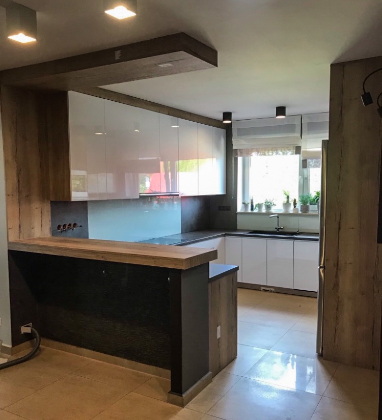 Nowoczesna kuchnia z białymi szafkami, drewnianymi akcentami i blatem barowym z widocznymi gniazdkami elektrycznymi.