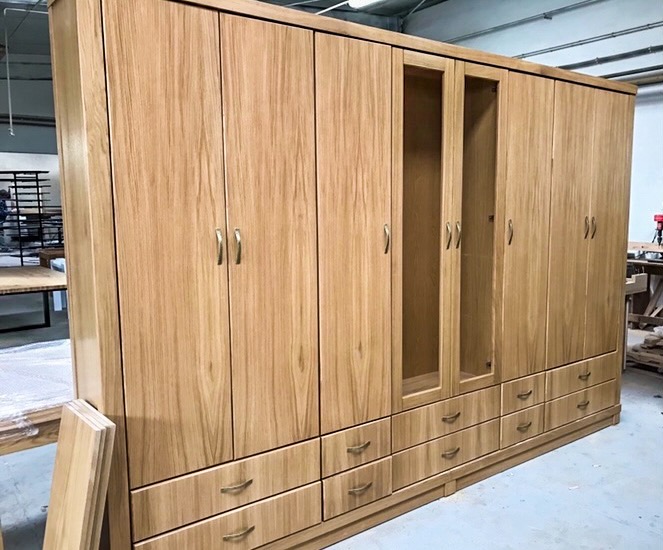 Duża, jasna szafa z drewna z licznymi drzwiczkami i szufladami, niektóre drzwiczki z przeszkleniami, złote uchwyty, widoczna struktura drewna.