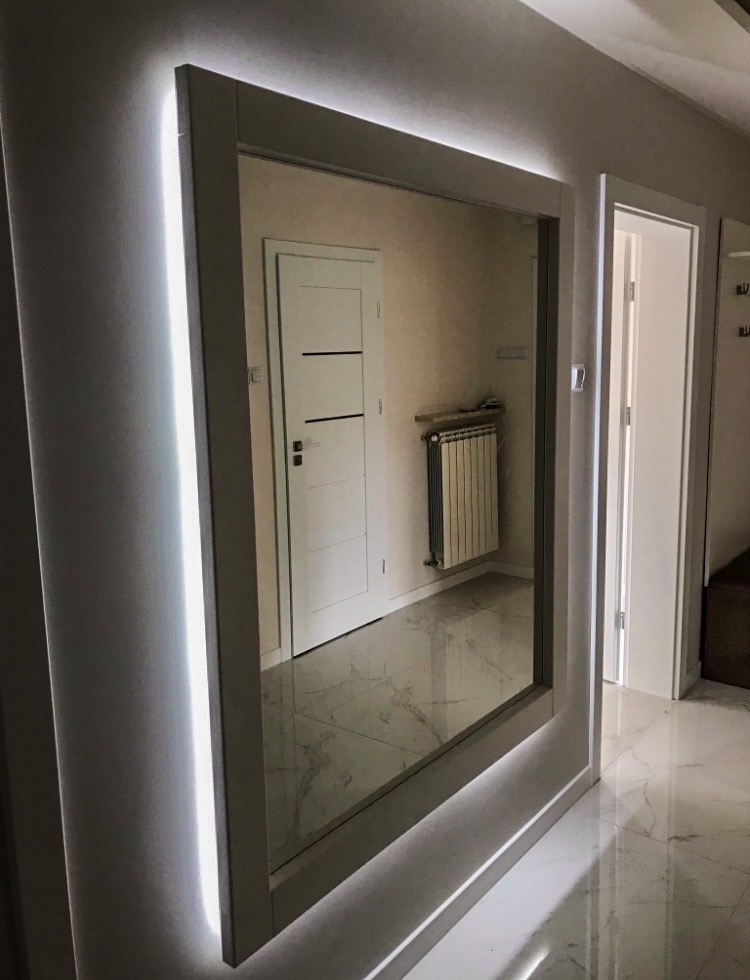 Nowoczesne lustro w ramie z podświetleniem LED, odbijające fragment przedpokoju z marmurową podłogą i białymi drzwiami.