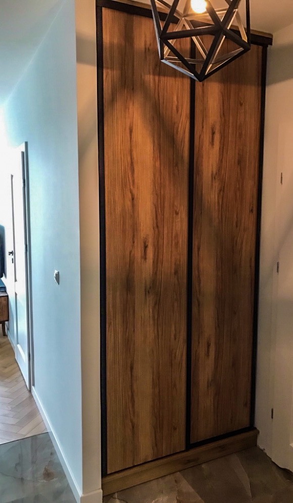 Dwudrzwiowa szafa przesuwna z frontami imitującymi drewno, z czarnymi obramowaniami, oświetlona lampą sufitową w kształcie geometrycznym, widoczna w korytarzu z jasnymi ścianami i parkietem.