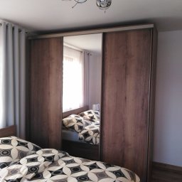 Kraina mebli Harasim karol - Szafa wnękowa z przesuwnymi drzwiami z lustrem, odbijająca fragment sypialni z łóżkiem w pościeli geometrycznej, oświetlona nowoczesną lampą sufitową.