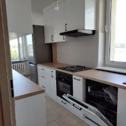 Nowoczesna kuchnia z białymi szafkami i drewnianym blatem, widoczna z otwartą zmywarką, okapem i płytą grzewczą.