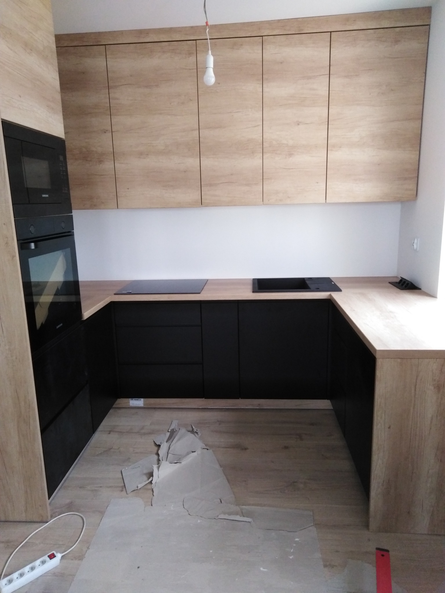 Niedokończona zabudowa kuchenna z czarnymi szafkami dolnymi i drewnopodobnymi górnymi, widoczne płyty indukcyjne, na podłodze kartony zabezpieczające.