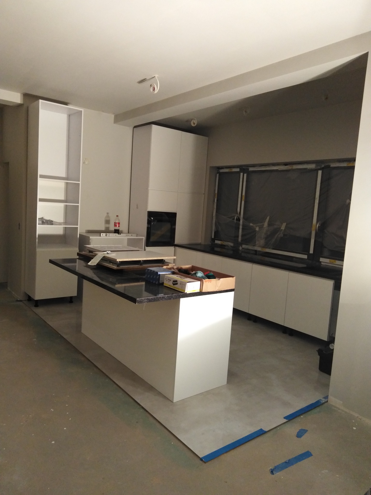 Wnętrze kuchni w trakcie montażu: białe szafki, wyspa z czarnym blatem, okno z folią ochronną, narzędzia i materiały na blacie wyspy.