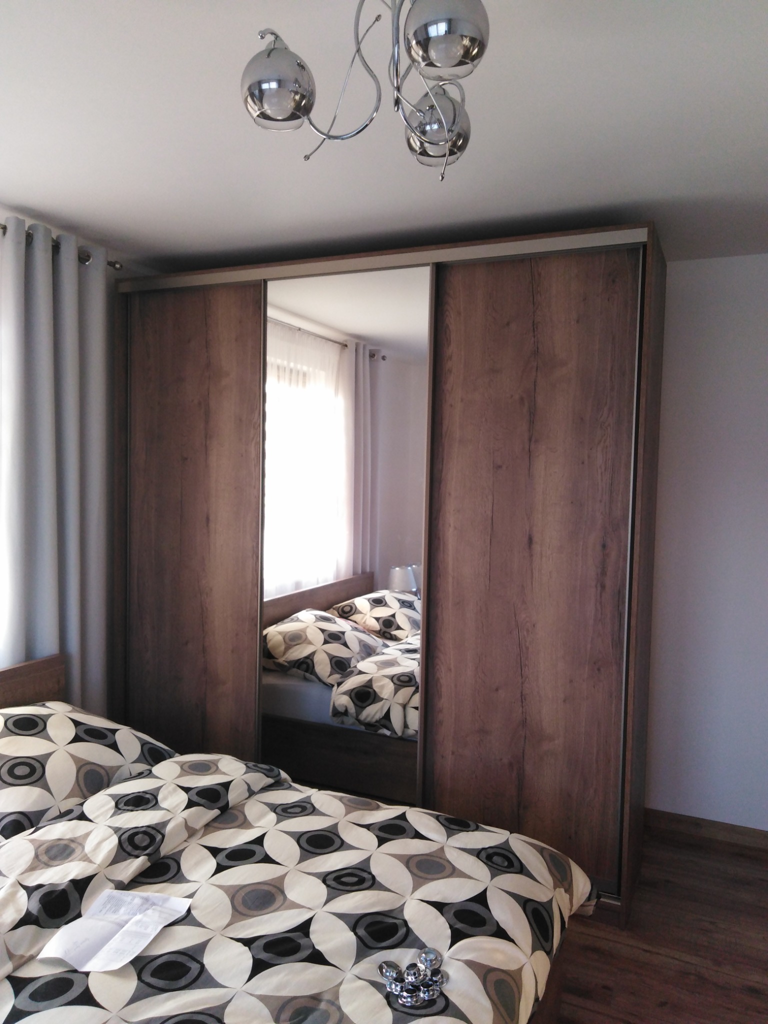 Szafa wnękowa z przesuwnymi drzwiami z lustrem, odbijająca fragment sypialni z łóżkiem w pościeli geometrycznej, oświetlona nowoczesną lampą sufitową.