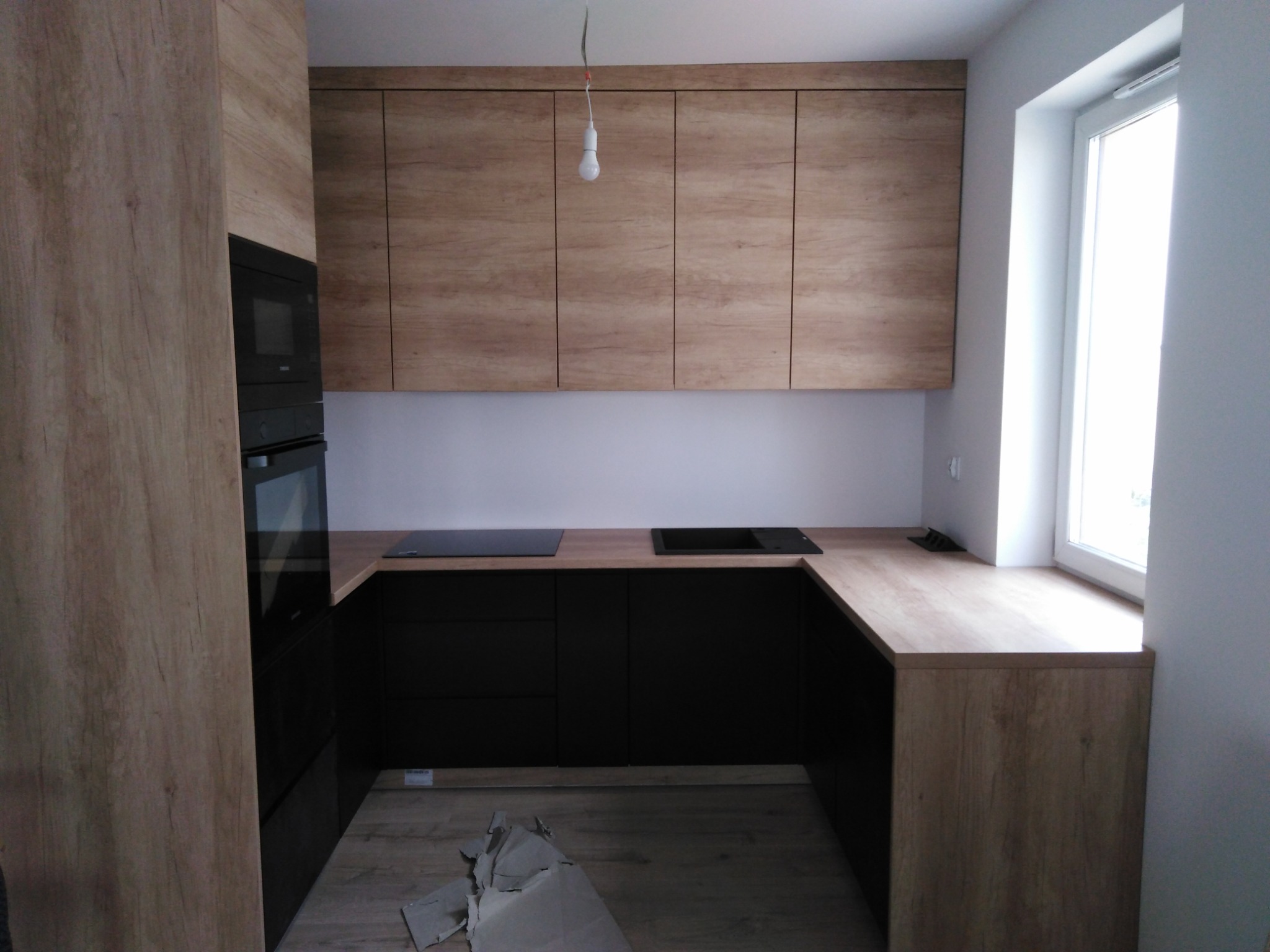 Nowoczesna kuchnia z czarnymi szafkami dolnymi i górnymi szafkami w odcieniach drewna, blat drewniany, płyta indukcyjna, piekarnik w zabudowie, widoczne elementy wykończeniowe i niedoskonałości...