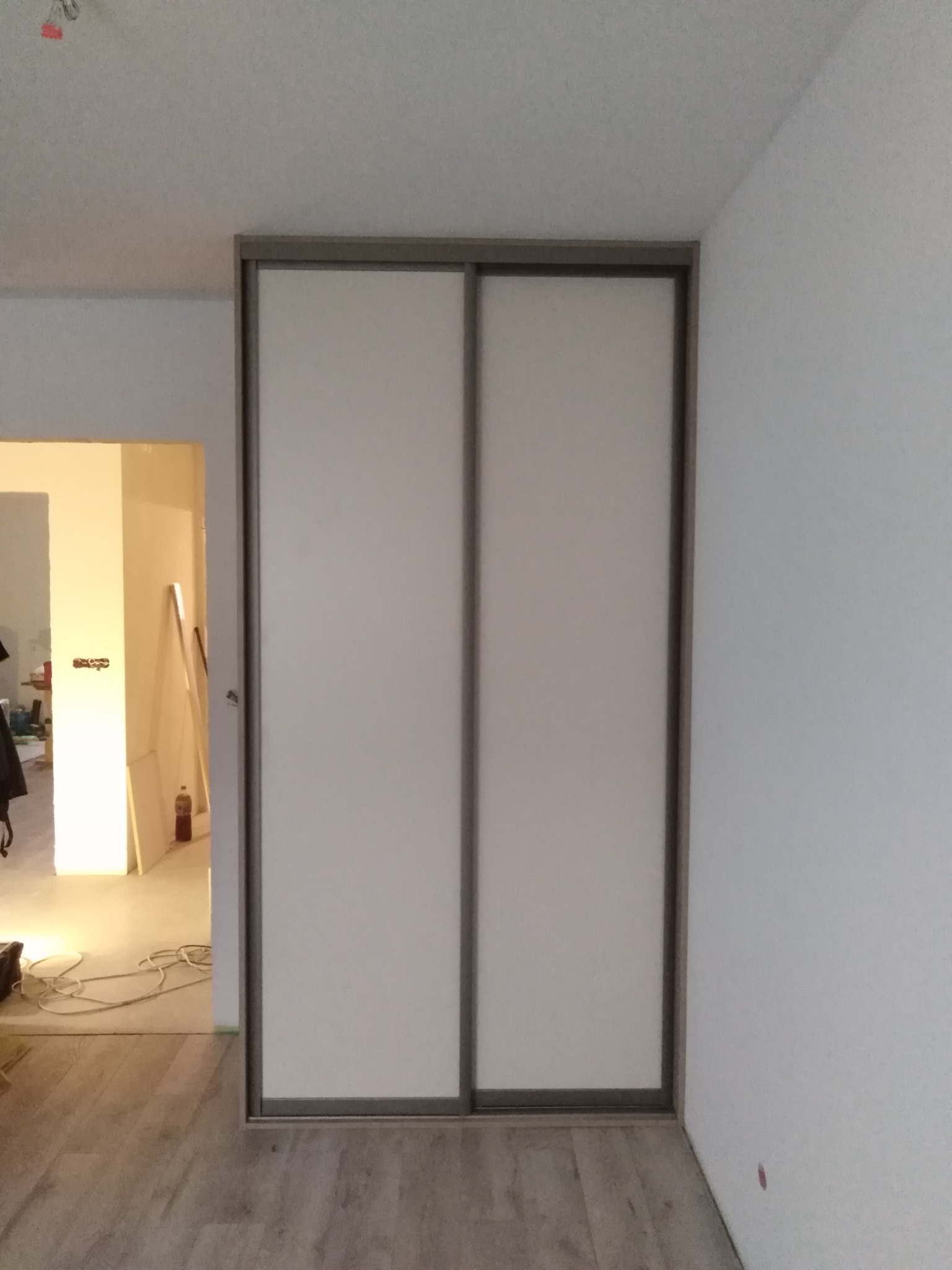 Zamknięta szafa z przesuwnymi, białymi drzwiami i szarą ramą w trakcie montażu, widoczne kable i narzędzia w tle, podłoga panelowa.