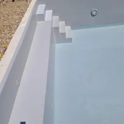 Wnętrze nowego basenu z białymi schodami i błękitną wodą, widoczne elementy oświetlenia podwodnego, otoczenie w trakcie wykończenia.