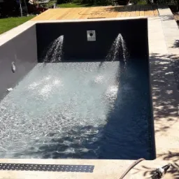 Nowoczesny basen z szarym wykończeniem i drewnianym tarasem, dwa strumienie wody wlewają się do basenu, tworząc efekt fontanny.